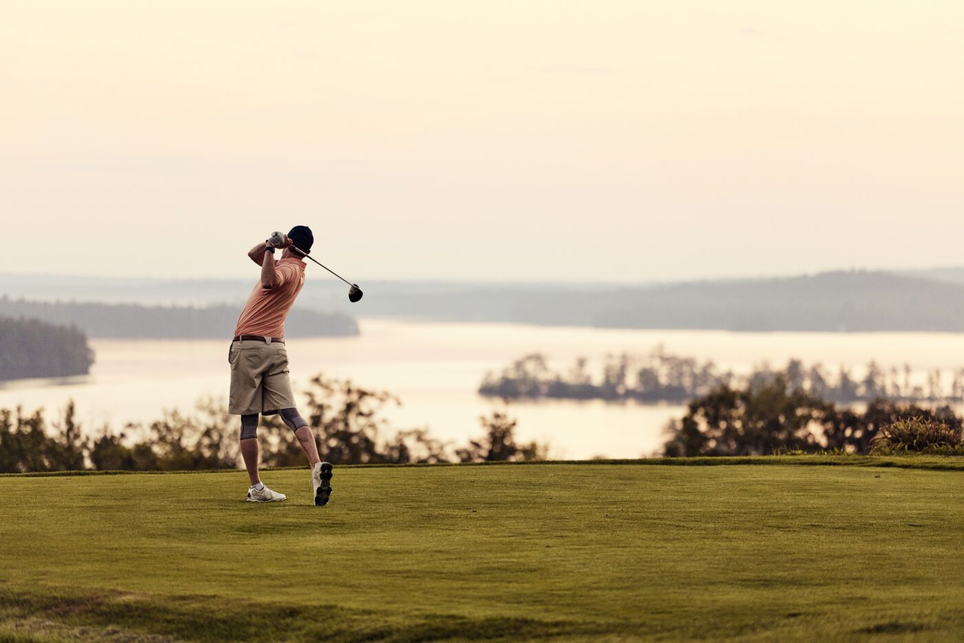 A person in shorts and a cap swings a golf club on a grassy course overlooking a lake and distant trees at sunset.