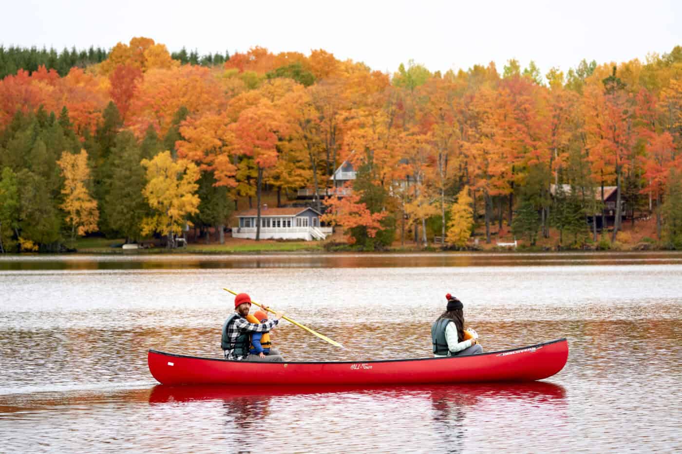 Two people paddle a red canoe on a calm lake with colorful autumn trees and lakeside houses in the background.