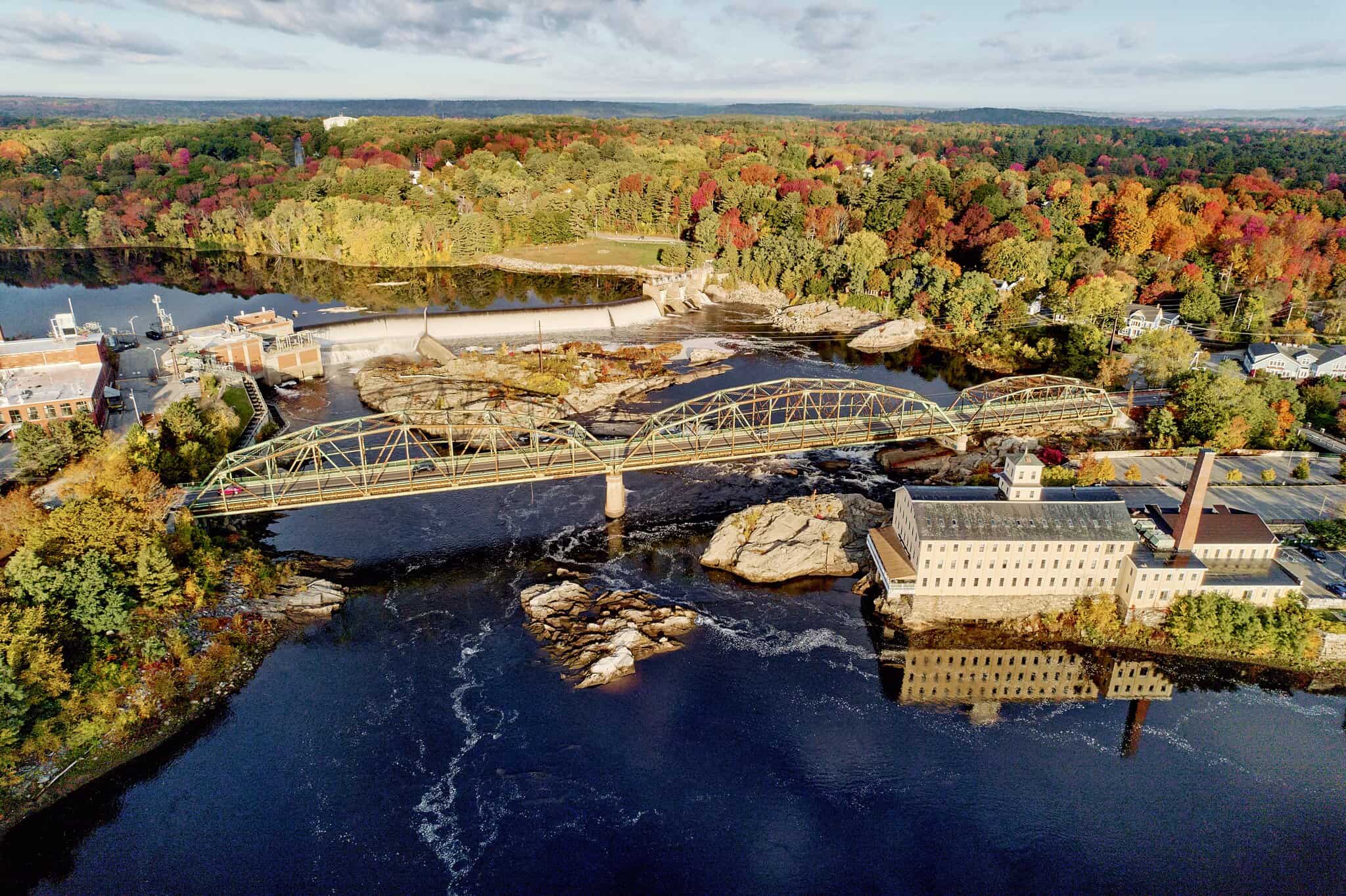 Aerial view of a bridge crossing a river with a dam and a large stone building surrounded by autumn trees.