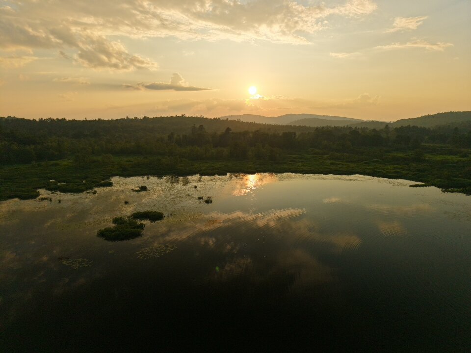 A calm lake reflects the sky and sun during sunset, with trees and distant hills in the background under partly cloudy skies.