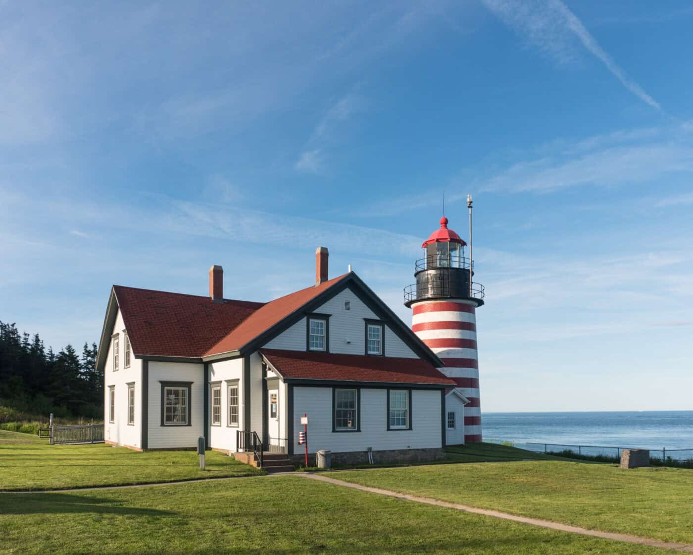 A white house with a red roof stands beside a red and white striped lighthouse on a grassy area by the ocean under a clear blue sky.