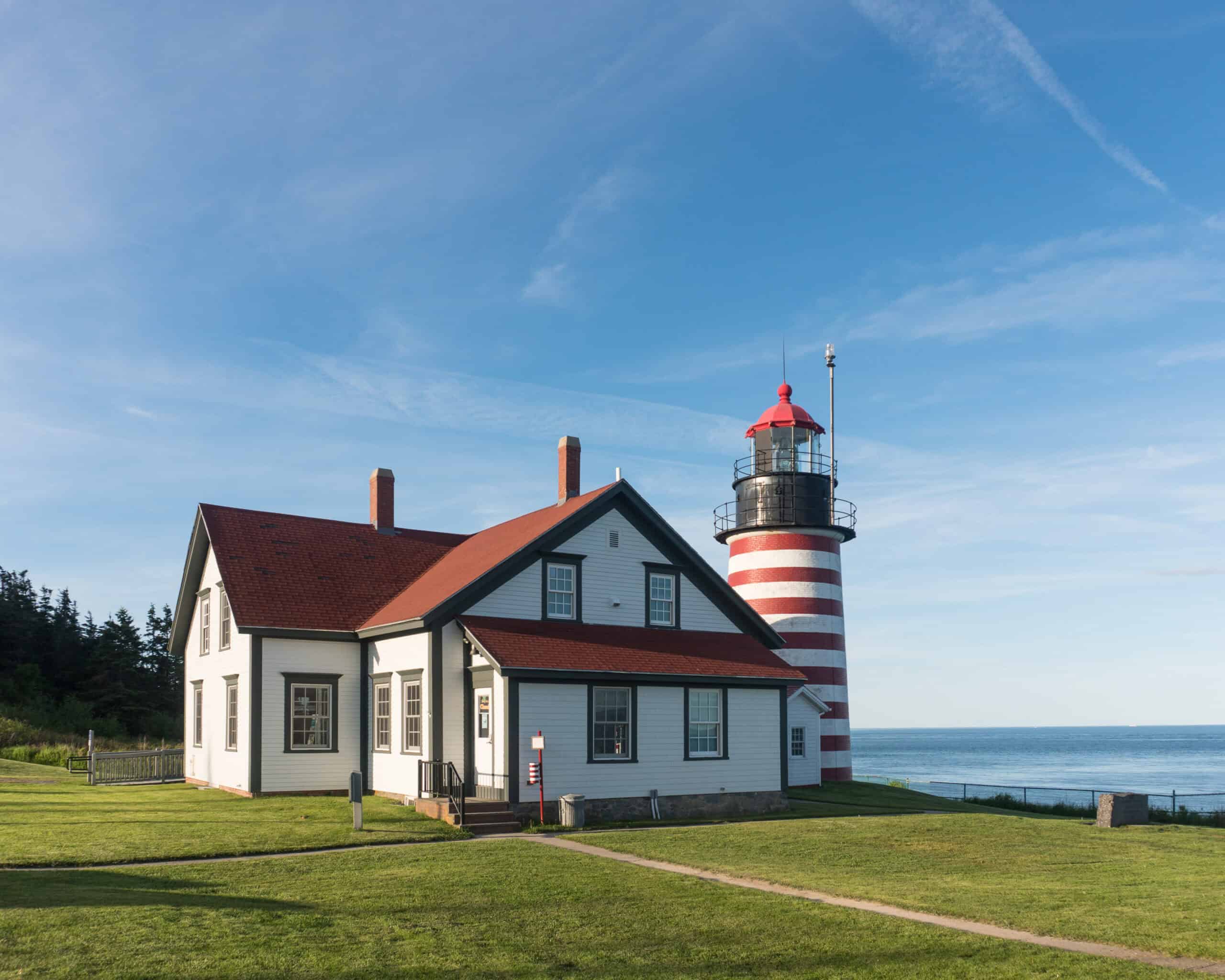 A white house with a red roof stands beside a red and white striped lighthouse on a grassy area by the ocean under a clear blue sky.