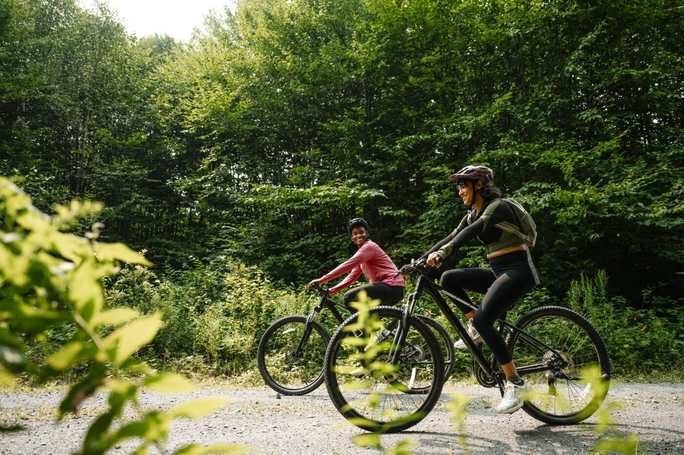 Two people wearing helmets ride bicycles on a gravel path surrounded by dense green foliage and trees on a sunny day.