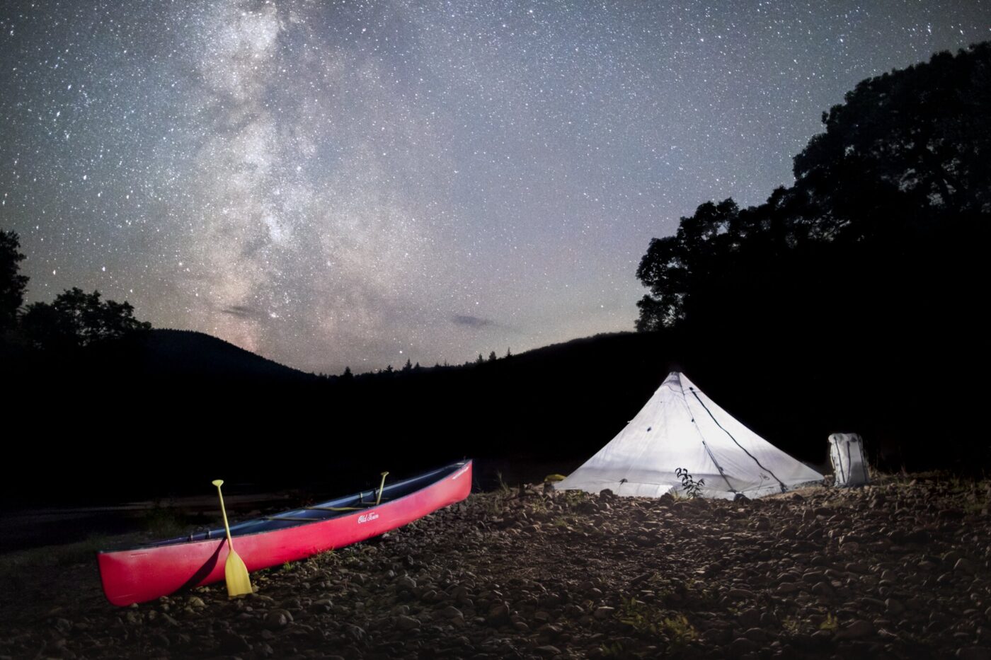A red canoe and a white tent are set up on rocky ground under a starry night sky with the Milky Way visible.