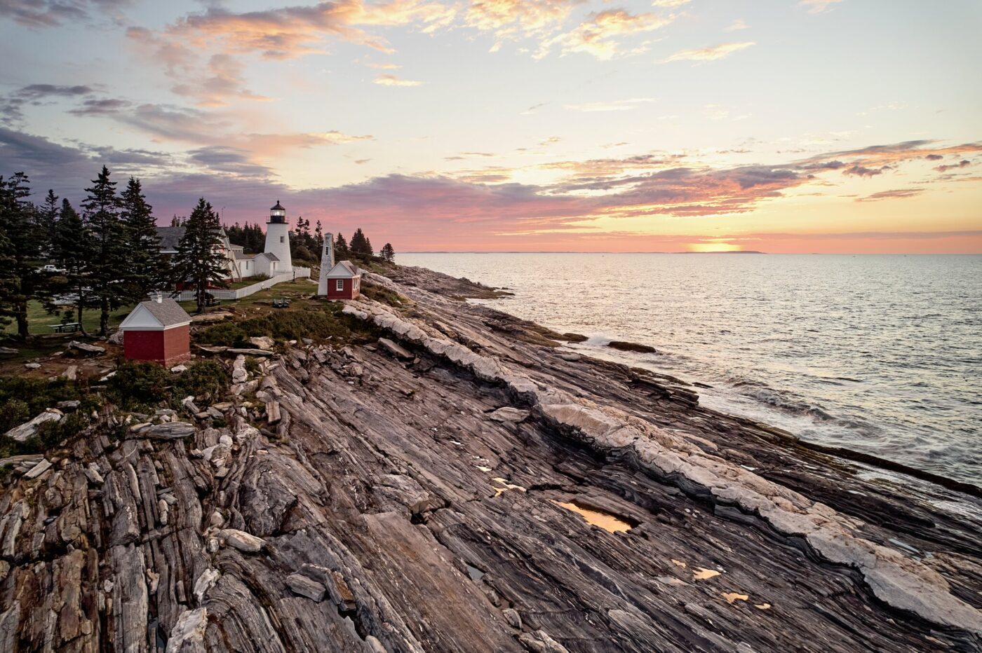 A lighthouse and small red buildings stand on rocky coastal cliffs at sunset, with the ocean and a partly cloudy sky in the background.