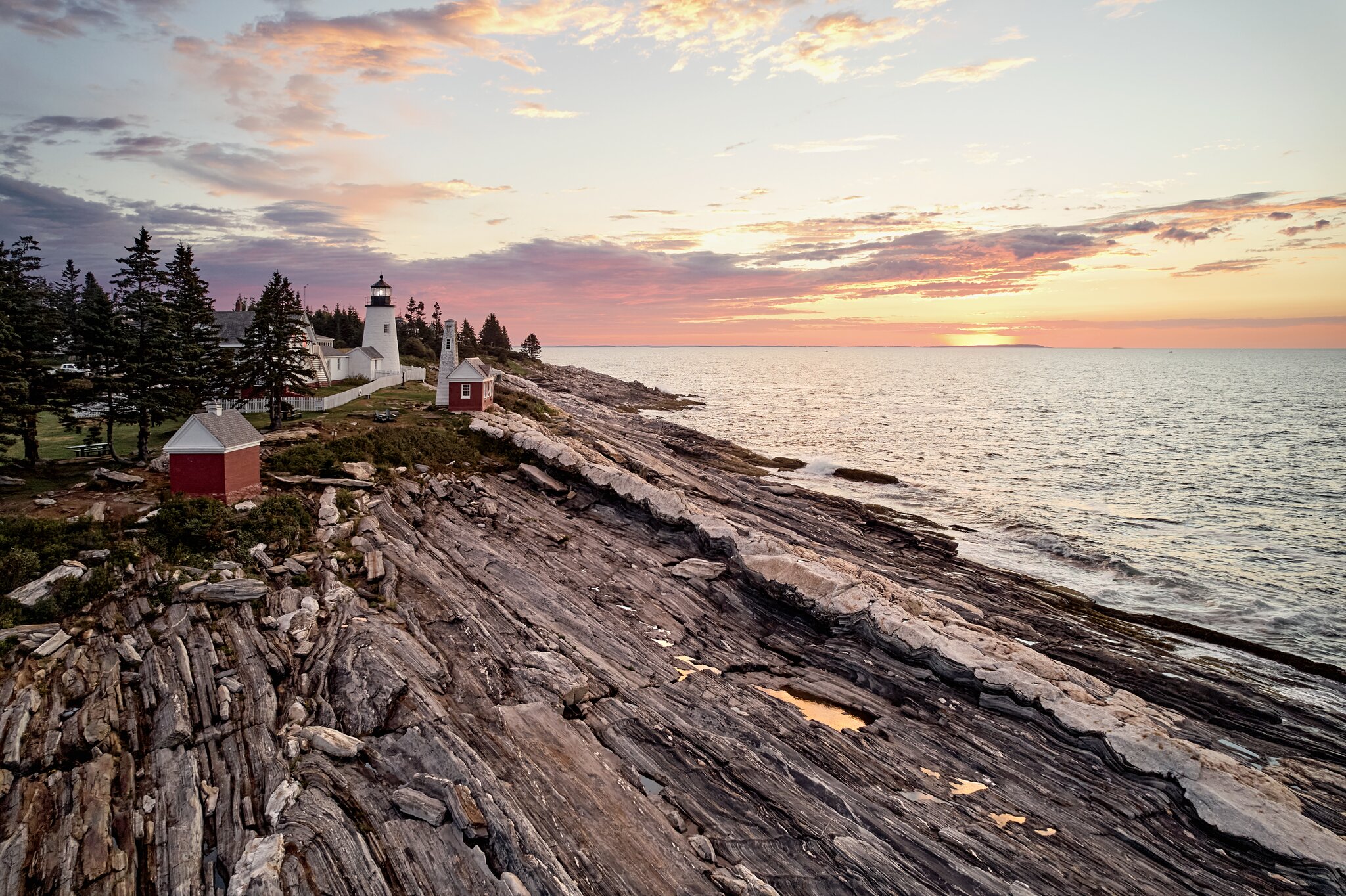 A lighthouse and small red buildings stand on rocky coastal cliffs at sunset, with the ocean and a partly cloudy sky in the background.