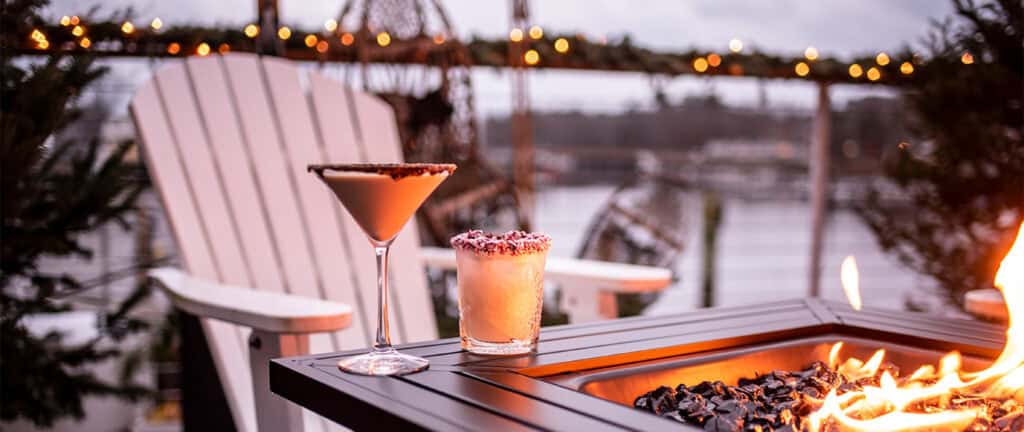 Cocktails placed on the edge of a lit fire pit next to a white Adirondack chair.