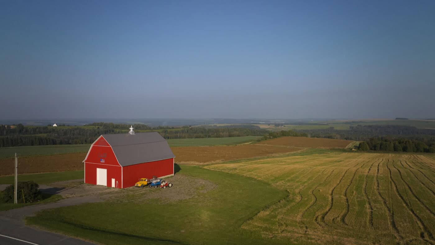 A red barn with white doors stands beside a mowed field and farmland under a clear sky, with tractors parked nearby.