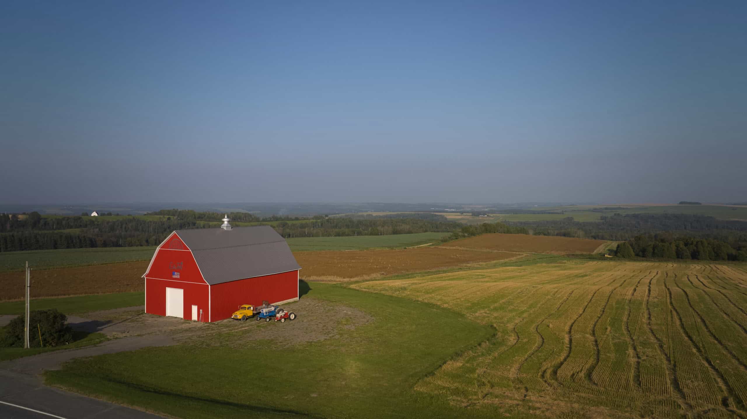 A red barn with white doors stands beside a mowed field and farmland under a clear sky, with tractors parked nearby.